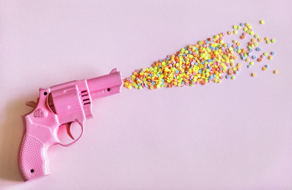 Photographie pop et colorée représentant un pistolet rose tirant des paillettes de plastiques multicolore, pouvant symboliser des bonbons ou des pilules chimiques.